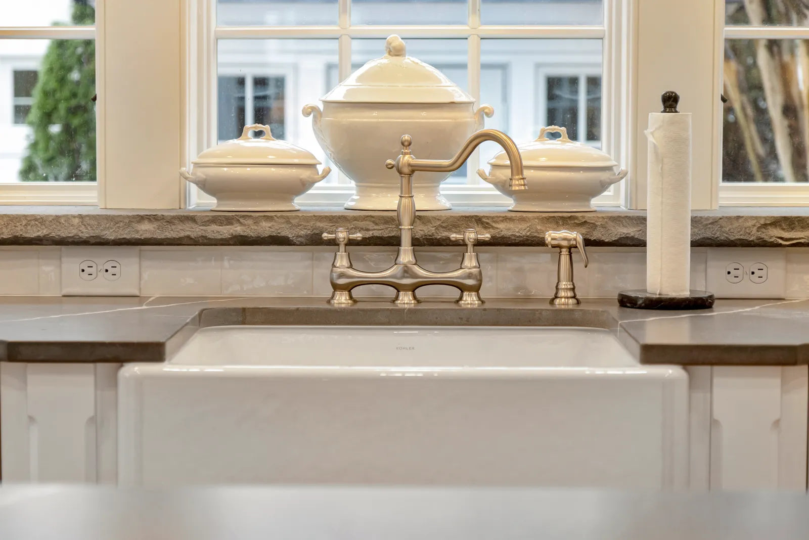 Polished nickel bridge faucet with twin handles mounted behind a white farmhouse sink in a classic kitchen