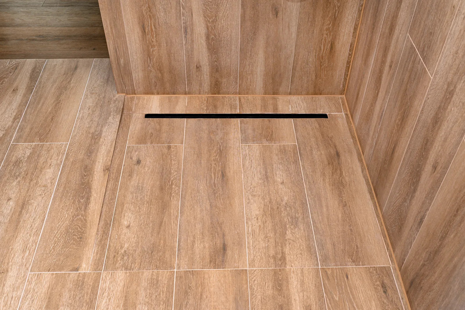 Stainless steel linear shower drain set into large-format grey tile along the wall of a modern walk-in shower