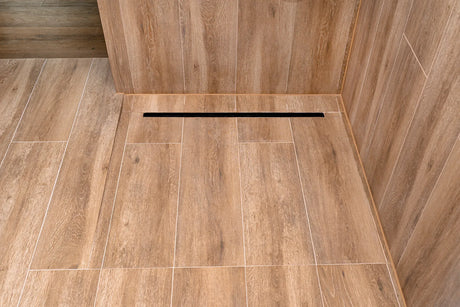 Stainless steel linear shower drain set into large-format grey tile along the wall of a modern walk-in shower