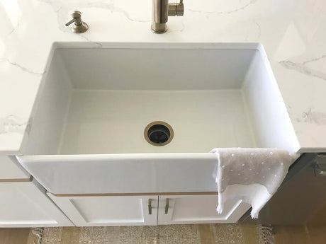 White fireclay farmhouse apron-front sink in a bright kitchen with a brass bridge faucet